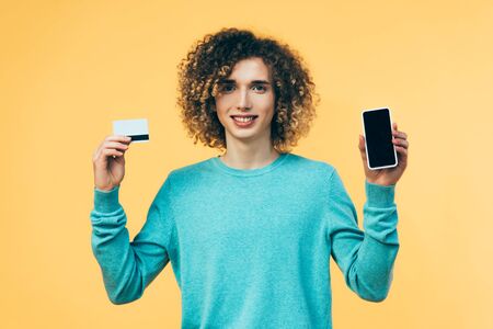smiling curly teenager holding smartphone and credit card isolated on yellow