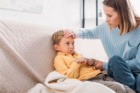 worried woman touching forehead of sick son sitting on sofaの写真素材