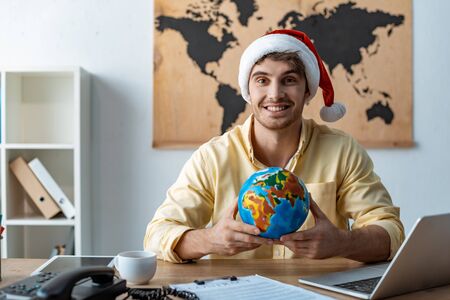 handsome travel agent in santa hat holding globe while smiling at cameraの写真素材