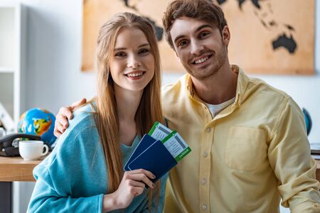 smiling man hugging girlfriend holding passports and air tickets in travel agencyの写真素材