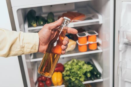 cropped view of man holding bottle of beer near open fridge full of foodの写真素材