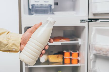 cropped view of man holding bottle of milk near open fridge with fresh food on shelves isolated on whiteの写真素材