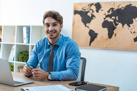handsome travel agent smiling at camera while sitting at workplaceの写真素材