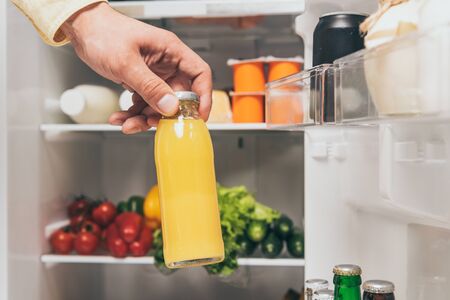 cropped view of man holding bottle of juice near open fridge with fresh food on shelvesの写真素材