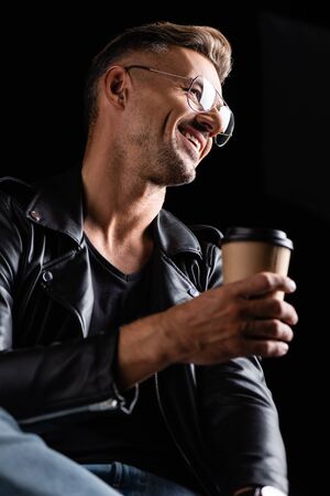 Low angle view of smiling man in sunglasses and leather jacket holding coffee to go isolated on blackの写真素材