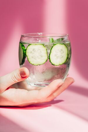 cropped view of woman holding glass of water with cucumber slices in sunlight on pink backgroundの写真素材