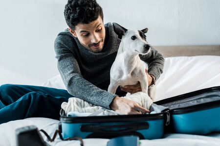 handsome and smiling bi-racial man packing luggage and holding jack russell terrierの写真素材
