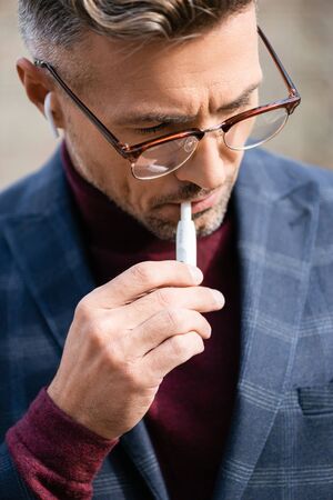 KYIV, UKRAINE - OCTOBER 11, 2019: Stylish businessman in eyeglasses and wireless earphones smoking iqosのeditorial素材