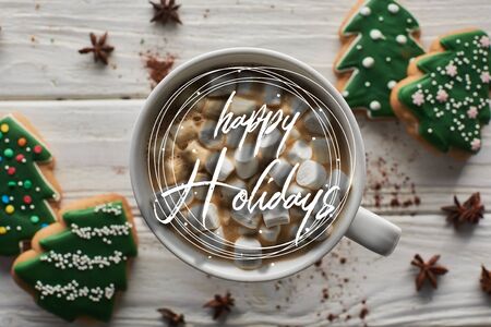 top view of Christmas cacao with marshmallow in mug on white wooden table near anise and cookies with happy holidays illustration の写真素材