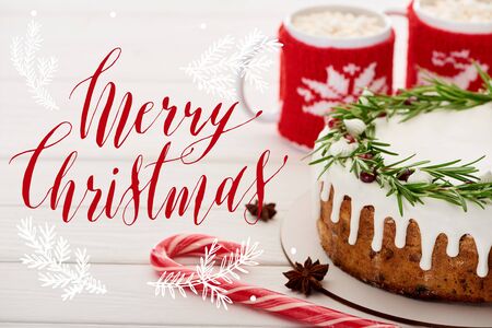 candy cane, christmas pie with icing and two cups of cocoa with marshmallows on white wooden table with Merry Christmas illustrationの写真素材