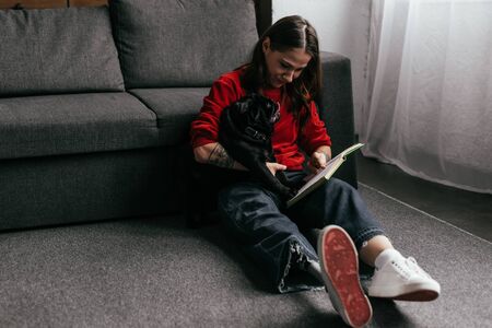 Woman with leg prosthesis reading book and holding pug dog on floor in living roomの写真素材