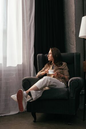 Girl with leg prosthesis holding coffee cup and looking at window in living room