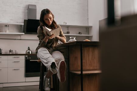 Selective focus of girl with prosthetic leg reading book by kitchen tableの写真素材
