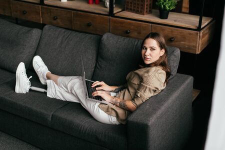 Young woman with prosthetic leg holding laptop and looking away on sofa
