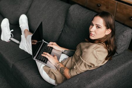 Beautiful girl with prosthetic leg using laptop with blank screen and looking away on sofa