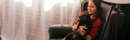 Smiling girl holding pug dog in armchair at home, panoramic shotの写真素材