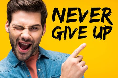 handsome man pointing with finger at never give up lettering and winking isolated on yellowの写真素材