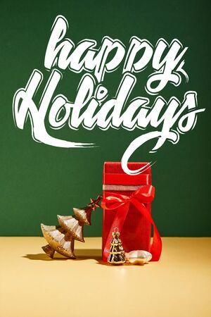 red gift box and decorative Christmas tree with golden baubles on green background with white happy holidays letteringの写真素材