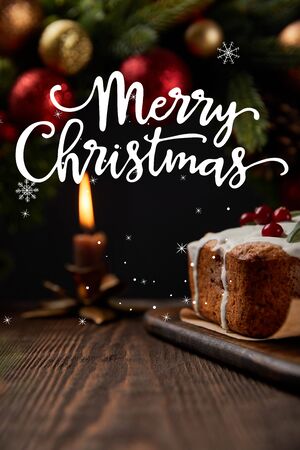 selective focus of traditional Christmas cake with cranberry near Christmas wreath with baubles and burning candle on wooden table with Merry Christmas illustrationの写真素材