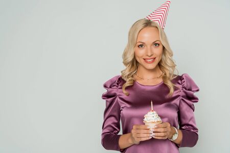 happy, elegant woman in party cap holding birthday cupcake and smiling at camera isolated on greyの写真素材