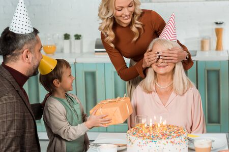 cheerful woman covering eyes of happy granny with hands, and smiling boy holding gift box