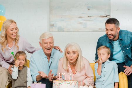 senior woman blowing out candles on birthday cake while sitting on sofa near happy familyの写真素材