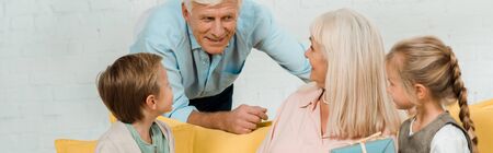 panoramic shot of senior man talking to happy wife sitting on sofa with grandchildren