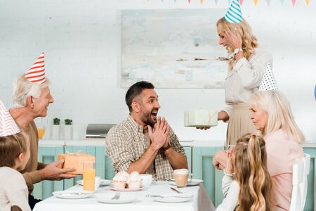 cheerful woman and senior man presenting gift boxes to excited man sitting at kitchen table near familyの写真素材