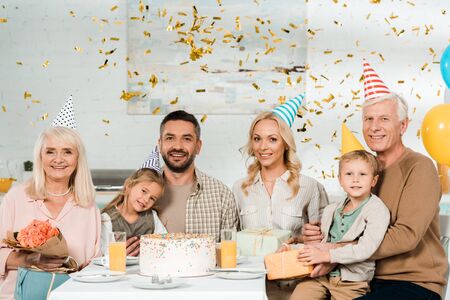 cheerful family sitting at kitchen table near birthday cake under falling confetti and smiling at cameraの写真素材