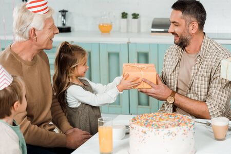 happy man taking gift box from daughter while sitting near birthday cake with familyの写真素材