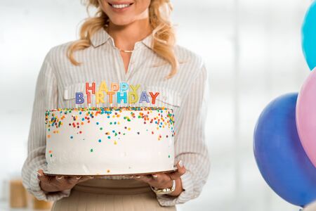 cropped view of smiling woman holding birthday cake with colorful candles and happy birthday letteringの写真素材