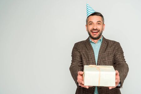 happy man in party cap holding gift box while looking at camera isolated on grey
