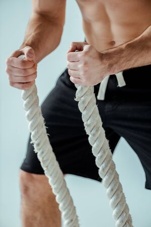 cropped view of sexy muscular bodybuilder with bare torso exercising with battle rope isolated on greyの写真素材