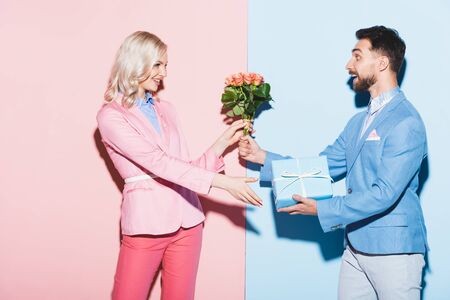 handsome man giving to smiling woman bouquet and gift on pink and blue backgroundの写真素材