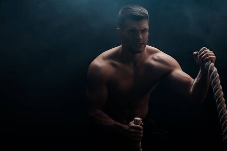 sexy muscular bodybuilder with bare torso excising with battle rope on black background with smokeの写真素材