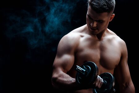 sexy muscular bodybuilder with bare torso excising with dumbbell on black with smokeの写真素材