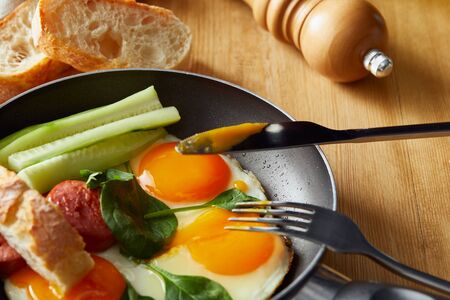 fried eggs in frying pan with vegetables and sausage near fork, knife, bread at wooden tableの写真素材