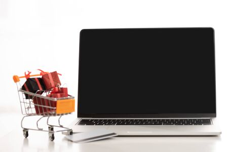 Laptop with blank screen and credit card with toy gifts in cart on table on white backgroundの写真素材