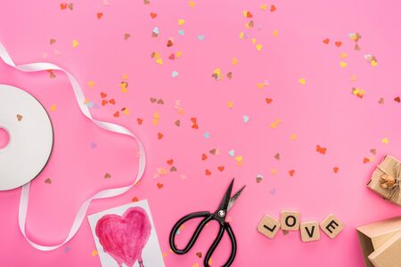 top view of valentines confetti, empty compact disk, scissors, gift boxes, greeting card and love lettering on wooden cubes on pink backgroundの写真素材