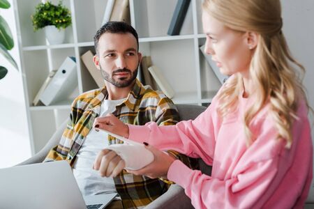 selective focus of caring woman putting bandage on injured hand of bearded man sitting with laptopの写真素材