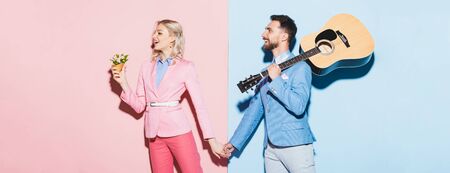 panoramic shot of shocked woman with bouquet and handsome man with acoustic guitar holding hands on...