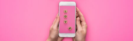 partial view of woman holding smartphone with image of cubes with love lettering on pink background, panoramic shotの写真素材