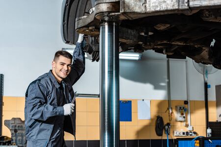 smiling mechanic holding wrench and looking at camera while standing under car raised on car liftの写真素材