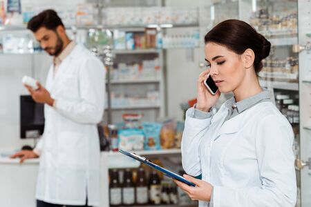 Selective focus of pharmacist with clipboard talking on smartphone with colleague at backgroundの写真素材