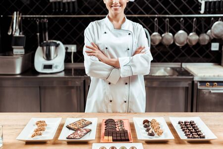 cropped view of happy chocolatier standing with crossed arms near tasty chocolate candies on platesの写真素材