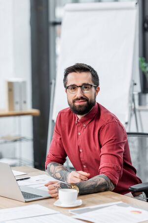 handsome and smiling account manager sitting at table and looking at camera