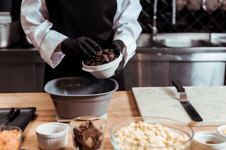 cropped view of chocolatier in latex gloves adding dark chocolate chips into bowl on kitchen scalesの写真素材
