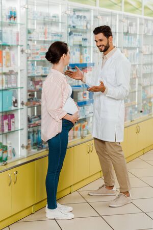 Smiling pharmacist looking at pregnant customer by medicaments on shelvesの写真素材