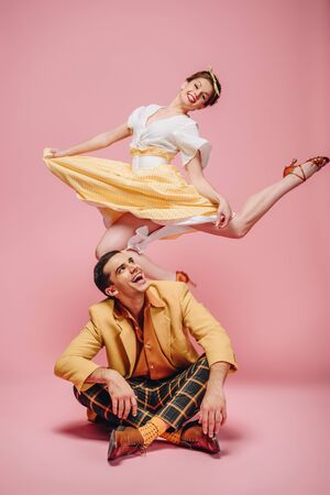 cheerful man sitting on floor and happy girl jumping while dancing boogie-woogie on pink backgroundの写真素材