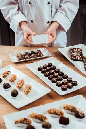 cropped view of chocolatier holding blank card near tasty chocolate candies on platesの写真素材